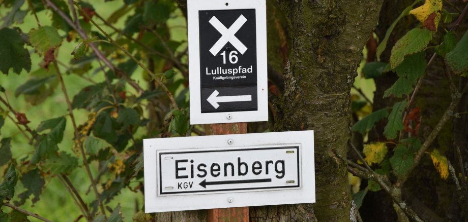 Zwei weiße Schilder an einem Holzpfeiler an einem Baum mit den Aufschriften: Eisenberg mit einem Pfeil, der nach links zeigt. Auf dem anderen Schild weiße Schrift auf schwarzem Hintergrund: "16 - Lulluspfad - Knüllgebirgsverein" mit einem linken Pfeil. Im Hintergrund Blätter von einem Baum.