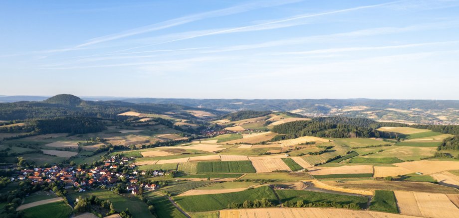 Im unteren Bildbereich ist eine Landschaft zu sehen. Neben einem kleinen Dorf mit einigen Häusern im linken Bereich sind vor allem Felder sichtbar. In der Landschaft gibt es einige Erhöhungen. Der Himmel ist blau und nur ganz leicht bewölkt.