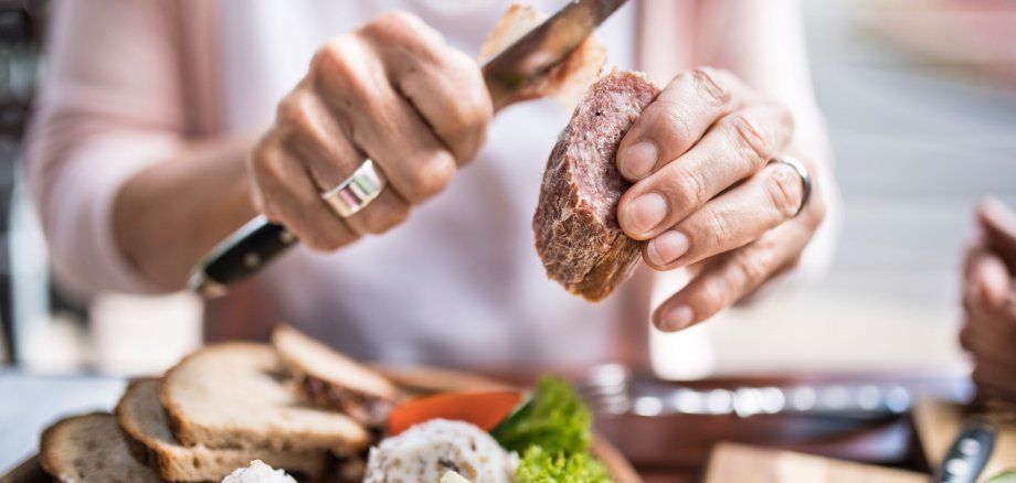 Eine Frau macht die Haut von einer Ahle Wurscht mit einem Messer ab. Man sieht einen Teller mit Wurst, Brot und sauren Gurken.