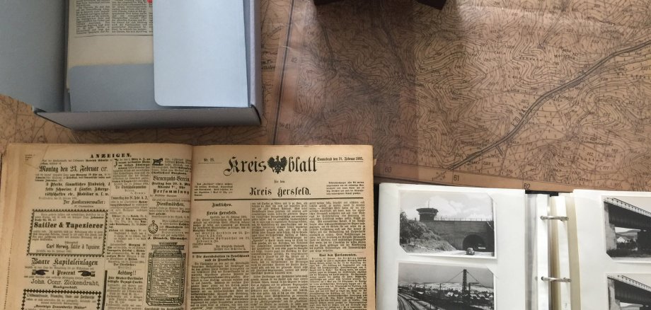 Links oben ein Karton, der halb geöffnet ist und worin alte Zeitungen liegen. Im Hintergrund liegt eine antike Karte, unten links eine historische Ausgabe des Kreisblatts von 1885. Rechts unten ein Fotoalbum mit schwarz-weiß Fotos vom Bahnhof Bebra und einem Tunnel. Oben in der Mitte liegt ein Dia, daneben steht Dorflinden.