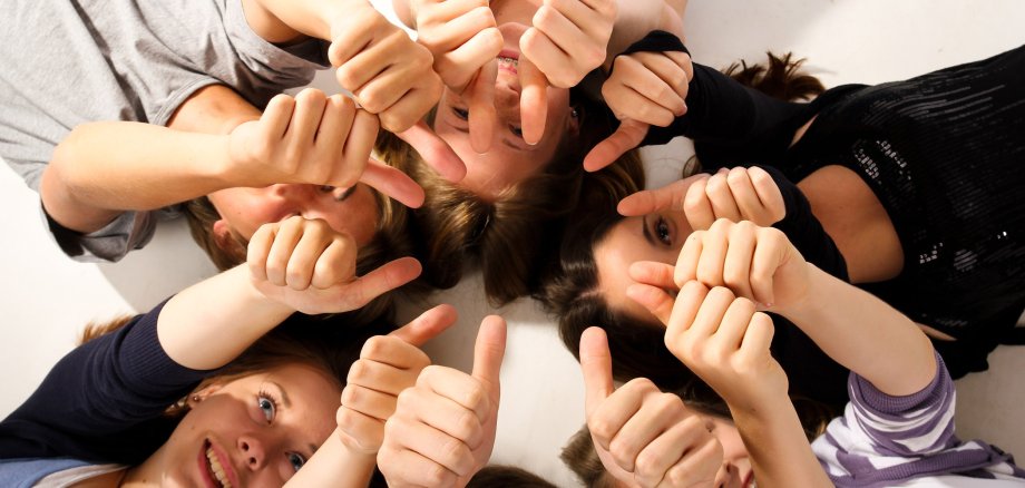 Sechs Kinder liegen auf dem Rücken, Kopf an Kopf in einem Kreis und strecken ihre Arme mit Daumen hoch aus.