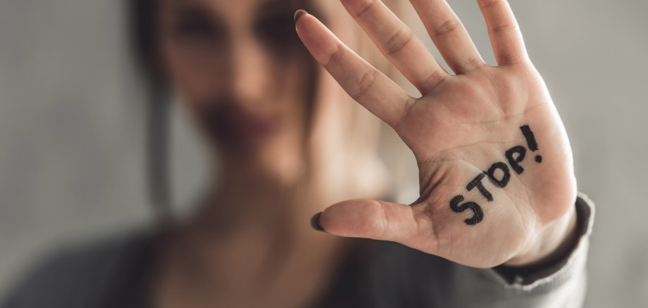 Im Hintergrund ein Portrait einer jungen Frau mit Blessuren im Gesicht. Im Vordergrund zeigt sie ihre Handinnenfläche, auf der in schwarzer Schrift "Stop!" steht. 