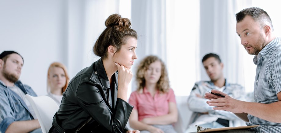 Rebellierende Teenagerin hört einem Sozialpädagogen zu während einer Gruppentherapiestunde mit anderen Jugendlichen, die Probleme haben.