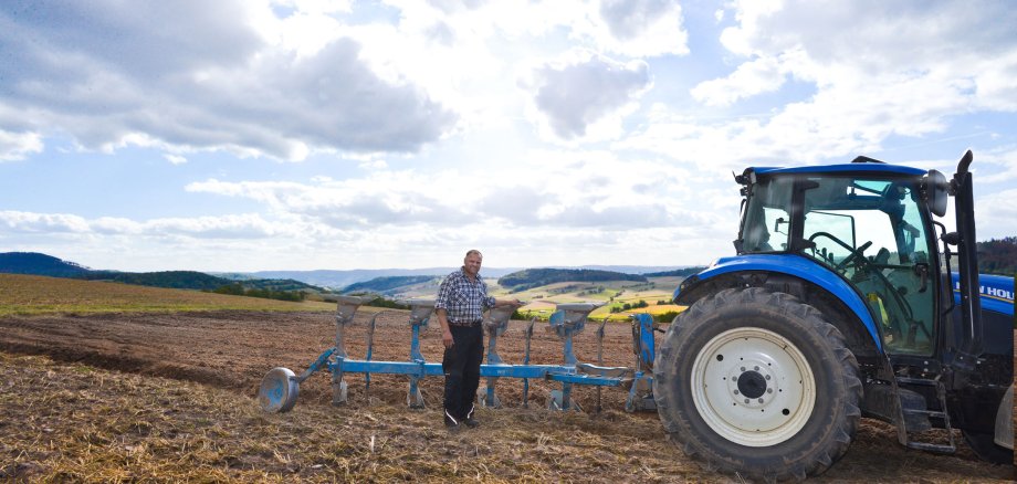 Ein Landwirt steht auf einem halb bearbeitetem Acker an seinem blauen Pflug, der am blauem Traktor hängt. Im Hintergrund ist eine Hügellandschaft mit Wald und Feldern sowie ein mit weißen Wolken halbbedeckter blauer Himmel zu sehen.