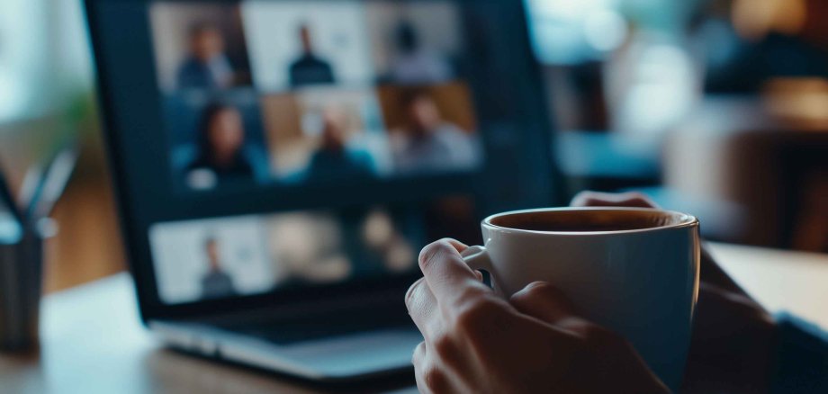 Virtual meeting concept with a close-up of hands holding a coffee mug in front of a laptop screen showing a video conference