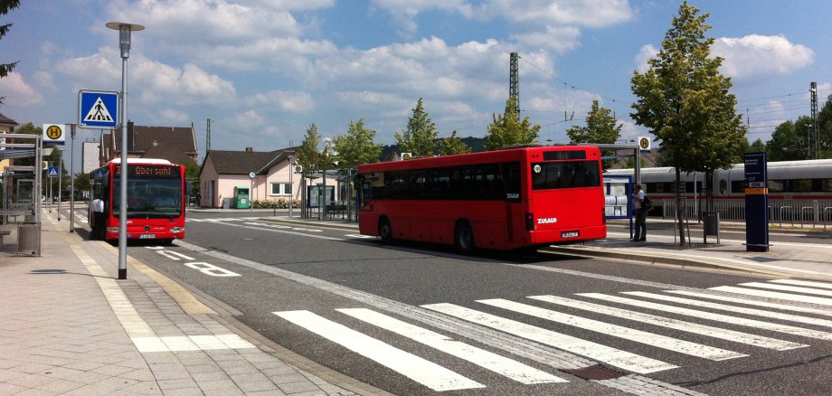 Bushaltestelle in Bad Hersfeld: Zwei rote Busse stehen an einer Bushaltestelle. Im Vordergrund ist ein Zebrastreifen. im Hintergrund das Bahnhofsgebäude sowie blauer Himmel mit Wolken.