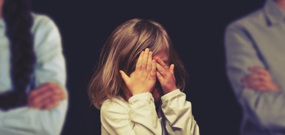 Kleines trauriges Mädchen hält sich die Hände vor das Gesicht. Im schwarzen Hintergrund sind rechts und links jeweils der Oberkörper einer Frau und eines Mannes zur Hälfte zu sehen. 