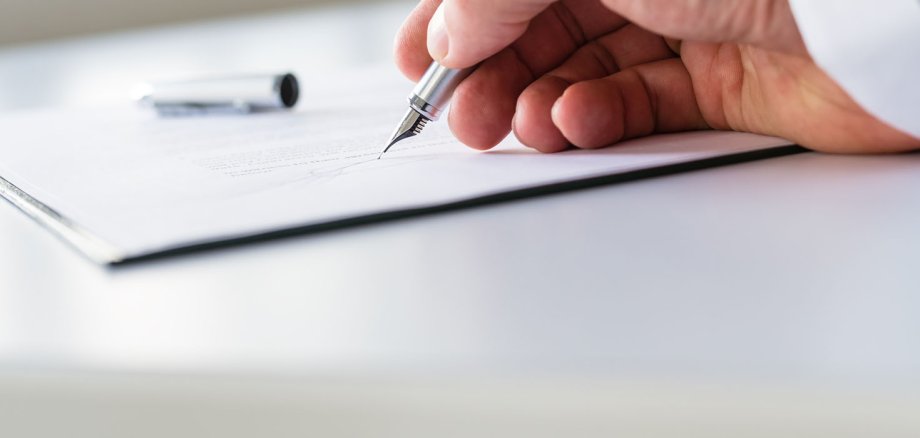 Eine männliche Hand unterschreib mit einem Füller ein Dokument. Das Dokument liegt auf einem Tisch. 