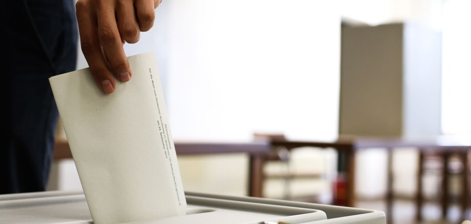 Eine männliche Hand wirft einen Wahlzettel in eine Wahlurne. Im Hintergrund ist eine Wahlkabine zu sehen.