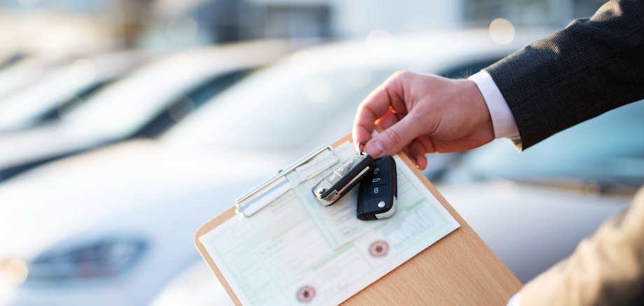 Die Hand eines Autoverkäufers halt einen Schlüssel und die Zulassungs-Papiere in der Hand. Im Hintergrund stehen Autos.