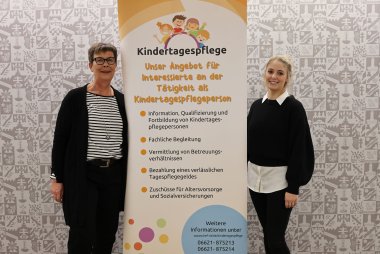 Zwei Personen stehen neben einem Banner mit der Aufschrift "Kindertagespflege". Sie lächeln in die Kamera. Im Hintergrund sieht man die Wappentapete des Landkreises Hersfeld-Rotenburg.