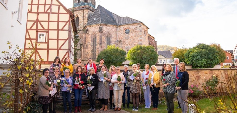 Eine Menschengruppe hauptsächlich aus geehrten Frauen, die jeweils eine Rose und Urkunde in der Hand halten, steht für ein Gruppenfoto vor einer Mauer in einem Garten. Im Hintergrund ist die Kirche von Rotenburg zu erkennen.