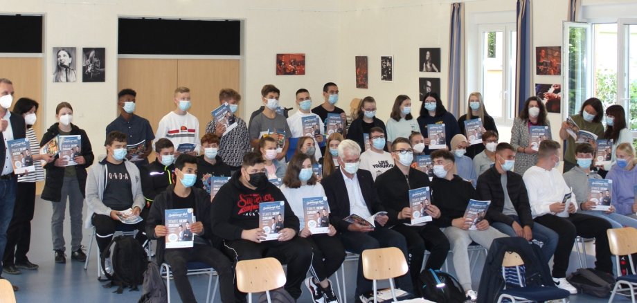 Eine Schulklasse sitzt mit Mundschutzmasken auf auf Stühlen bzw. die letzte Reihe steht und hält eine blaue Broschüre mit der Aufschrift "Ausbildungs-Navi - starte deine Zukunft" hoch. 