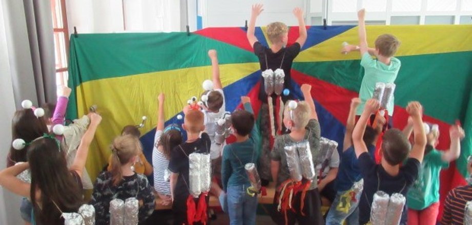 Rückansicht einer Kindergruppe, die alle eine silberne gebastelte Rakete auf ihren Rücken tragen. Die Kids strecken die Hände, als ob sie auf den Mars reisen. Sie stehen vor einer bunten Wand. 