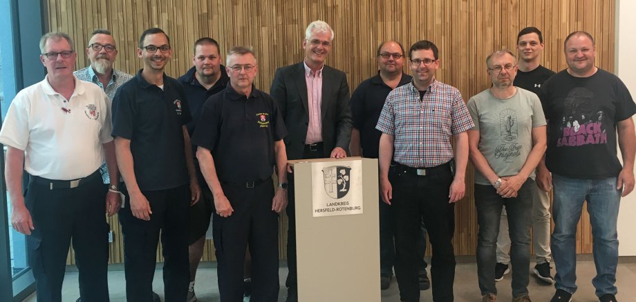 11 Männer stehen an einem Pult, auf dem ein Wappen des Landkreises Hersfeld-Rotenburg abgebildet ist. Sie schauen in die Kamera und lächeln. Einige der Männer tragen ein Feuerwehr-T-Shirt. In der Mitte steht Landrat Torsten Warnecke.