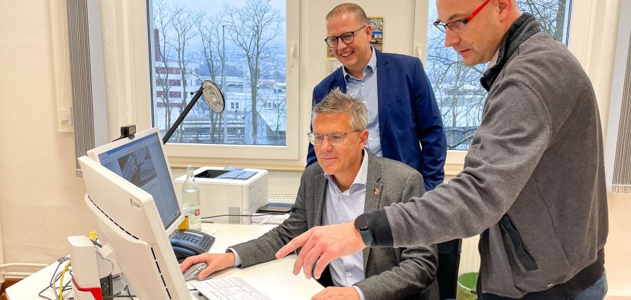 Erster Kreisbeigeordneter Dirk Noll sitzt an einem Schreibtisch und guckt auf zwei Bildschirme. Hinter ihm stehen zwei Mitarbeiter aus dem Landkreis und zeigen auf den Bildschirm.