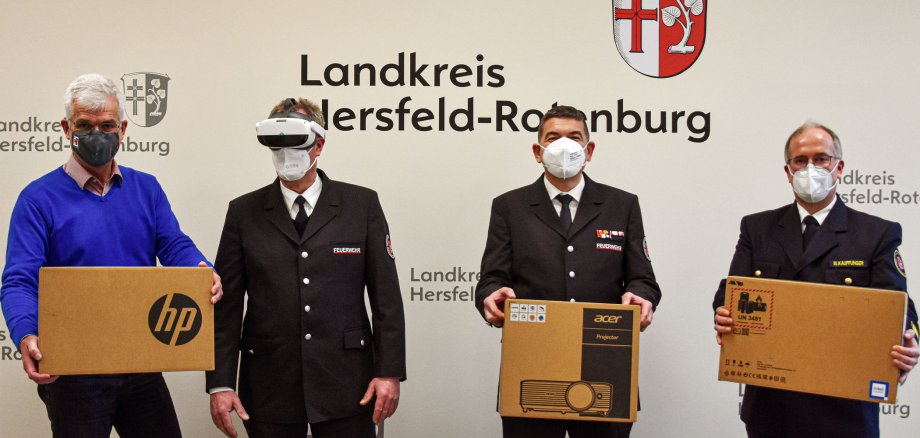 Vier Männer mit Schutzmaske stehen vor einer Wand mit dem Wappen und Schriftzug Landkreis Hersfeld-Rotenburg- Drei der Männer halten Kartons der Marken HP, Acer und Intel hoch. Ein Mann hat eine virtuelle Brille.