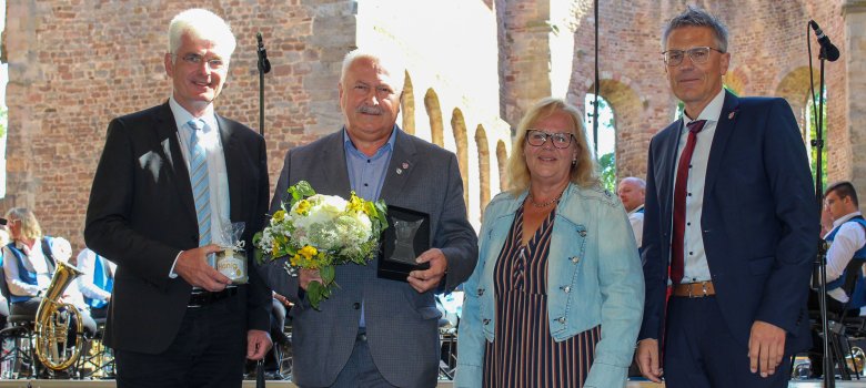 Landrat Warnecke, Alt-Bürgermeister Preßmann, Kreistagsvorsitzende Petra Wiesenberg und Erster Kreisbeigeordneter Dirk Noll stehen auf einer Bühne in der Stiftsruine in Bad Hersfeld. Preßmann hält einen Blumenstrauß und eine Ehrenplakette. Im Hintergrund sind Teile der Bergmannskapelle Hattorf zu sehen.