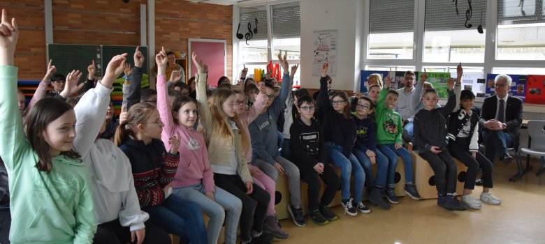 Die Schülerinnen und Schüler der vierten Klassen der Kreuzbergschule sitzen in ihrem Musikraum. Einige von ihnen melden sich und wollen eine Frage beantworten. Am Bildrand sitzt Landrat Torsten Warnecke.