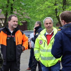 Ein Mann in einer orangefarbenen Jacke mit der Aufschrift "HessenForst" steht mit anderen Einsatzkräften in einem ländlichen Bereich.