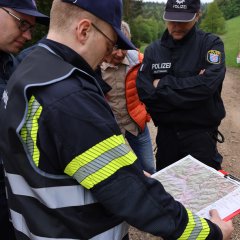 Ein Feuerwehrmann in reflektierender Schutzkleidung steht zusammen mit anderen Einsatzkräften und schaut auf eine Karte. Die Gruppe steht auf einem Feldweg.