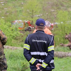 Ein Soldat in Tarnkleidung und ein Mann in einer reflektierenden Weste mit "Übungsleitung" im Waldgebiet. Mehrere Feuerwehrfahrzeuge stehen in der Nähe.