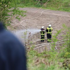 In der Ferne stehen zwei Feuerwehrleute im Wald und üben das Löschen mit dem Feuerwehrschlauch.