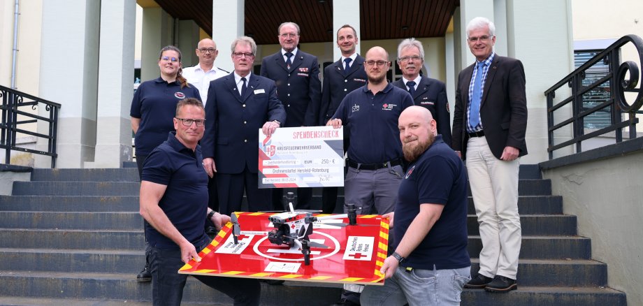 Mehrere Menschen stehen auf der Treppe vor dem Landratsamt in Bad Hersfeld. Einige tragen eine Feuerwehruniform. Die beiden Personen in der ersten Reihe halten einen Drohnenlandeplatz und eine Drohne. Die beiden Personen dahinter halten einen Spendenscheck. Die Personen lächeln in die Kamera.