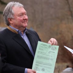 Ein älterer Mann mit grauem Haar und dunklem Mantel hält ein grünes Dokument in der Hand und lächelt, während er zur Seite blickt. Er trägt ein kariertes Hemd und einen Blazer.