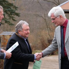 Zwei Männer geben sich die Hand. Der eine trägt einen karierten Anzug und eine rote Krawatte, der andere einen dunklen Mantel und ein grünes Dokument. Im Hintergrund ist eine Holzhütte zu sehen.