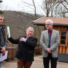 Drei Männer stehen draußen auf einer Holzterrasse. Ein Mann in der Mitte gestikuliert mit einem grünen Dokument, während die anderen beiden zuhören. Ein vierter Mann steht im Hintergrund.