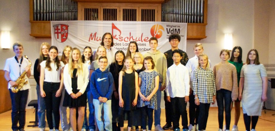 Das Bild zeigt eine Gruppe von Schülern und einen Lehrer der Musikschule des Landkreises Hersfeld-Rotenburg, die nach einem Konzert auf einer Bühne stehen. Ein Schüler hält ein Saxophon, und im Hintergrund ist ein Banner sowie eine große Orgel zu sehen.