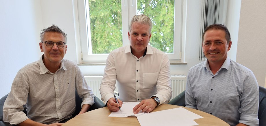 Drei Männer sitzen an einem runden Tisch in einem hellen Büro. Im Hintergrund sieht man große Fenster. Der Mann in der Mitte trägt ein weißes Hemd und unterschreibt einen Vertrag. Alle schauen in die Kamera.