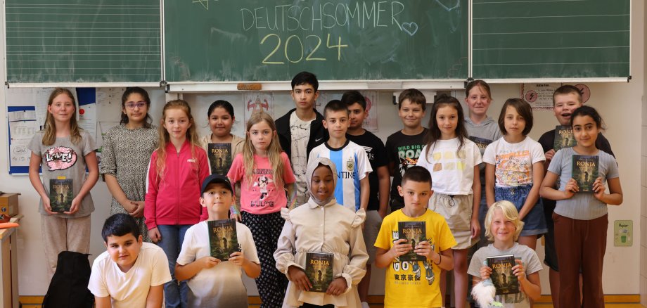 Rund 25 Schülerinnen und Schüler stehen vor einer Tafel in einem Klassenraum. Auf der Tafel steht mit Kreide geschrieben: "Herzlich willkommen im Deutschsommer 2024". Die meisten Kinder halten eine Ausgabe des Buches Ronja Räubertochter in der Hand.