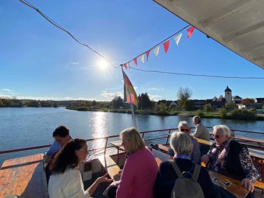 Mehrere ältere Personen sitzen an Deck eines Schiffes. Im Hintergrund sieht man Wasser und blauen Himmel mit strahlender Sonne.