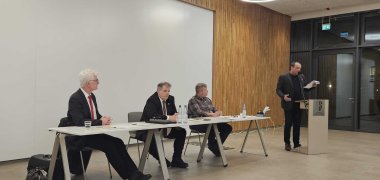 Ein Mann steht an einem Rednerpult im Konferenzraum und hält einen Vortrag. Er gestikuliert mit einem Blatt Papier in der Hand. Vor ihm sitzen drei Männer an einem langen Tisch, hören aufmerksam zu und haben Wasserflaschen und Unterlagen vor sich.