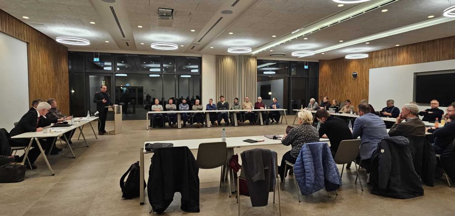 Ein großer Konferenzraum mit Holzverkleidung und modernen Lichtkreisen an der Decke. Zahlreiche Teilnehmende, darunter Landwirte, Kommunalvertreter und Kreisvertreter, sitzen an langen Tischen in einer U-Form und verfolgen eine Diskussion. Eine Person steht am Rednerpult und spricht zu den Anwesenden, während einige Zuhörende Notizen machen.