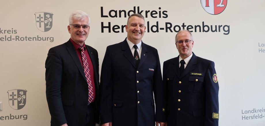 Drei Männer stehen vor einer Tapete mit der Aufschrift "Landkreis Hersfeld-Rotenburg". Zwei von ihnen tragen eine Feuerwehruniform. Der Mann in der Mitte hält eine Urkunde und eine Stracke.