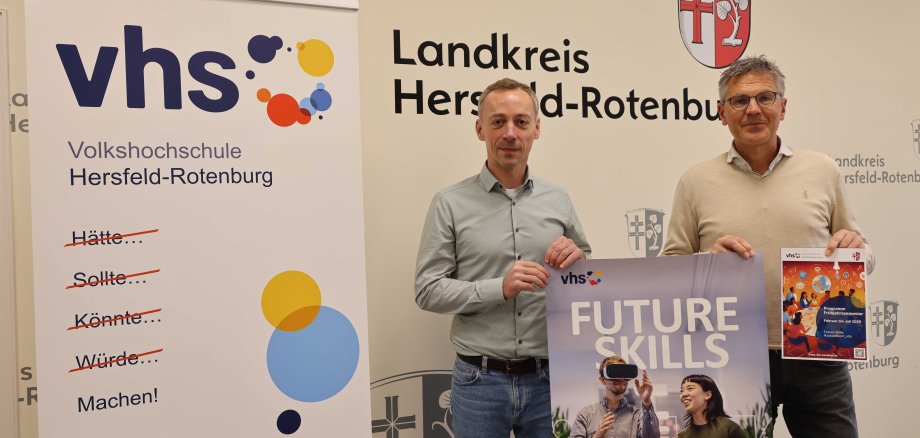 Fabian Göbel, Leiter der Volkshochschule, und Dirk Noll, Erster Kreisbeigeordneter, stehen vor einem Banner der Volkshochschule (VHS) Hersfeld-Rotenburg und einem Schild mit dem Wappen und Schriftzug des Landkreises Hersfeld-Rotenburg. Fabian Göbel hält ein Poster mit der Aufschrift „Future Skills“ und einer Abbildung von Personen mit VR-Brille. Dirk Noll präsentiert das Programmheft der VHS für das Frühjahrsemester 2025. Das Banner trägt den Slogan „Hätte... Sollte... Könnte... Würde... Machen!“ und zeigt bunte Kreise als Gestaltungselemente.