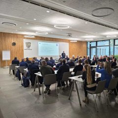 Ein großer Konferenzraum mit mehreren Reihen von Teilnehmern, die an Tischen sitzen und auf einen Vortragenden vorne blicken. An der Wand wird eine Präsentation mit dem Titel „Ausgangslage“ projiziert, die verschiedene Themen anspricht. Der Raum hat moderne Einrichtung mit Holzverkleidung an der Vorderwand, einer Uhr und dem Wappen des Landkreises Hersfeld-Rotenburg.