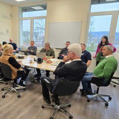 Ein Besprechungsraum mit mehreren Personen, die in einer Runde an einem Tisch sitzen und diskutieren. Auf dem Tisch stehen Getränke und Snacks, während einige Teilnehmer Notizen machen. An den Wänden sind das Wappen und der Schriftzug des Landkreises Hersfeld-Rotenburg zu sehen.