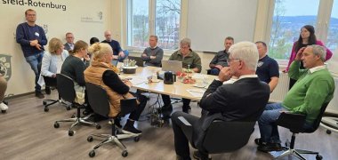 Ein Besprechungsraum mit mehreren Personen, die in einer Runde an einem Tisch sitzen und diskutieren. Auf dem Tisch stehen Getränke und Snacks, während einige Teilnehmer Notizen machen. An den Wänden sind das Wappen und der Schriftzug des Landkreises Hersfeld-Rotenburg zu sehen.