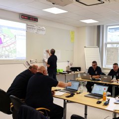 Mehrere Personen sitzen in einem Schulungsraum an Tischen, die mit Laptops ausgestattet sind, und arbeiten konzentriert. Im Hintergrund schreibt eine Person an einer Tafel, während links eine Karte auf eine Leinwand projiziert wird. Die Wände zeigen Notizen und Flipcharts mit Informationen.