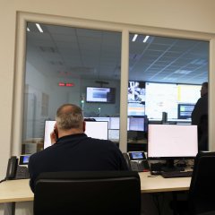 Ein Blick durch eine Glaswand in einen Überwachungsraum, in dem Personen an Computern arbeiten. Auf Bildschirmen und an der Wand werden Daten und Karten angezeigt, während im Hintergrund weitere Personen stehen und diskutieren.