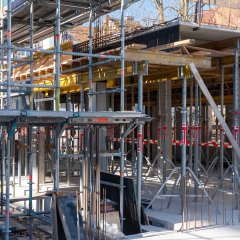 Die Baustelle mit Baugerüsten, Stahlträgern und Betonwänden ist im Hintergrund sichtbar. Metallverstrebungen ragen aus dem Beton heraus.