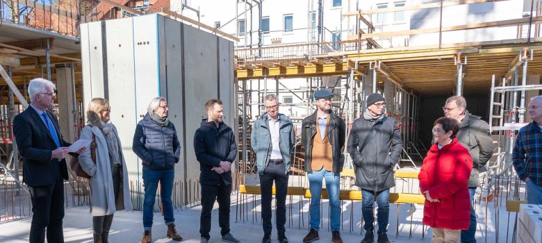 Eine Gruppe von neun Personen steht in einer Reihe auf der Baustelle. Im Hintergrund sind Baugerüste und der Rohbau des entstehenden Gebäudes zu sehen.